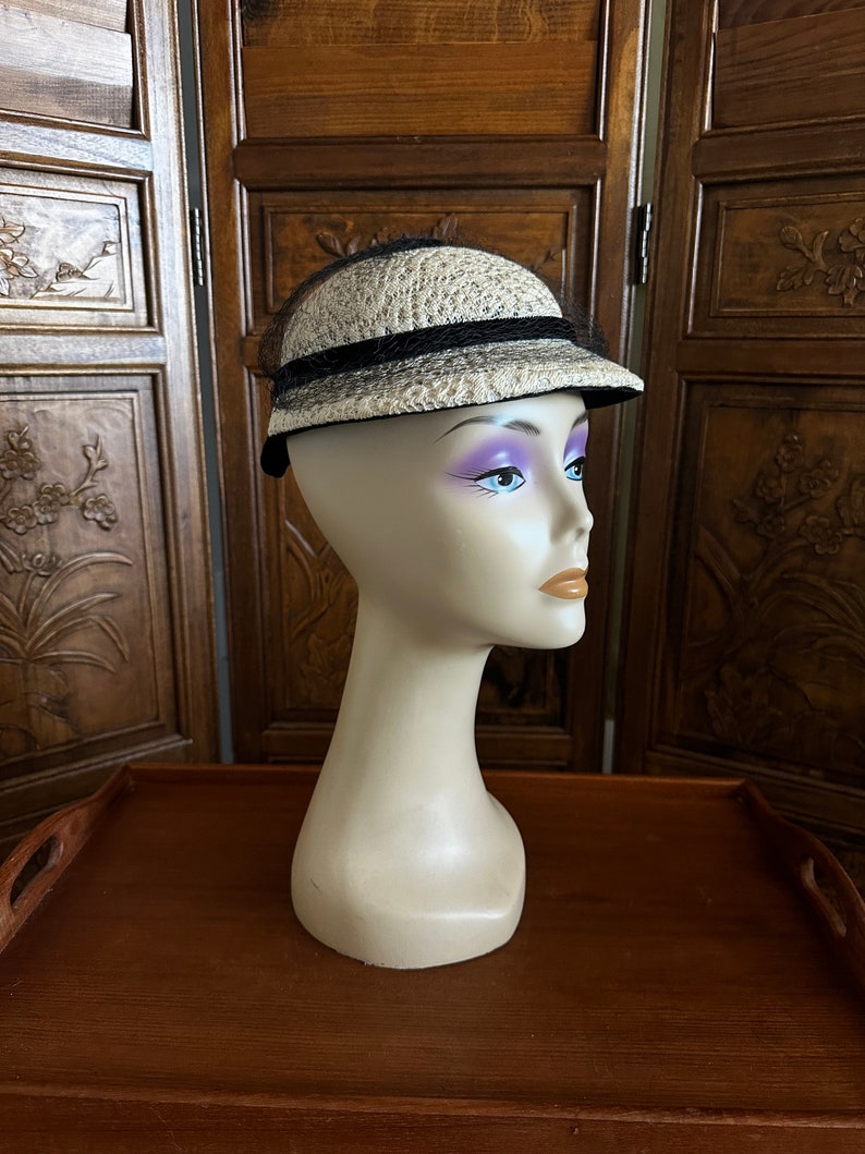 May include: A white straw hat with a black ribbon and black netting. The hat is on a mannequin head.