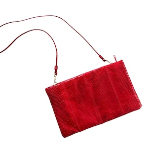 Pode incluir: Uma bolsa tiracolo retangular vermelha com um padrão texturizado de pele de cobra. A bolsa inclui uma alça de ombro fina e vermelha, fixada com ferragens metálicas douradas, e um fecho de correr ao longo de toda a parte superior.