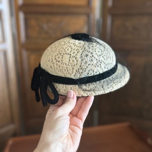 May include: A white lace hat with a black velvet band and bow. The hat has a rounded shape and a slightly curved brim.