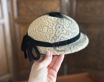 May include: A white lace hat with a black velvet band and bow. The hat has a rounded shape and a slightly curved brim.