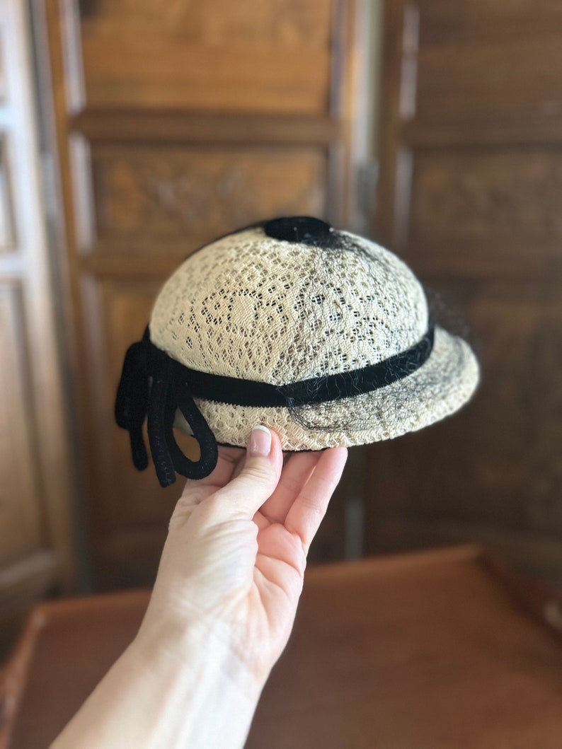 May include: A white lace hat with a black velvet band and bow. The hat has a rounded shape and a slightly curved brim.