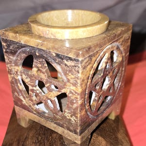 May include: A square brown stone oil burner with a carved pentagram design. The burner has a small, round well in the center for oil and a base with four legs.