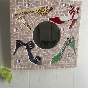 May include: A square mosaic mirror with a round mirror in the center. The mosaic is made of small tiles in various colors, including red, green, blue, and brown. The tiles are arranged in a pattern of shoes, with a red shoe, a green shoe, a blue shoe, and a brown shoe.