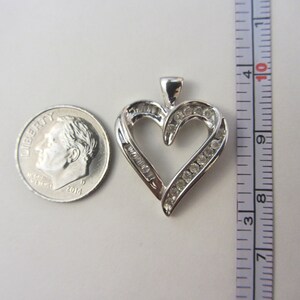 May include: A silver heart-shaped pendant with clear gemstones. The pendant is approximately 1 inch in height. A U.S. dime is next to the pendant for size comparison. The pendant has a loop at the top for a chain.