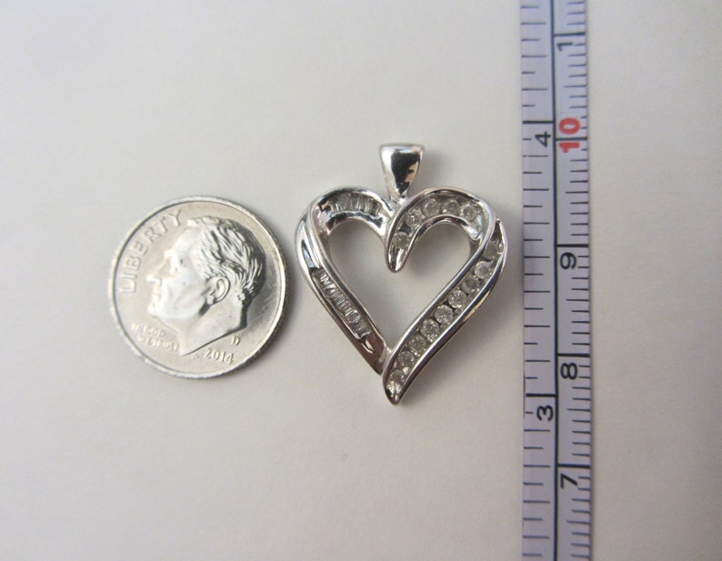 May include: A silver heart-shaped pendant with clear gemstones. The pendant is approximately 1 inch in height. A U.S. dime is next to the pendant for size comparison. The pendant has a loop at the top for a chain.