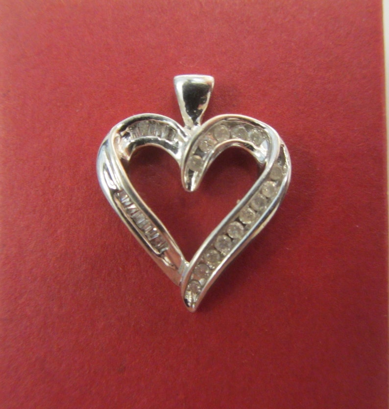 May include: A silver heart-shaped pendant with a bail at the top. The pendant is encrusted with small, round, and baguette-cut gemstones. The heart is set against a red background.