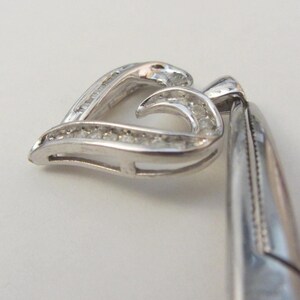 May include: A silver pendant in the shape of a heart, encrusted with small, clear gemstones. The pendant is shown at an angle, with the heart shape slightly open. The metal has a polished, reflective surface.