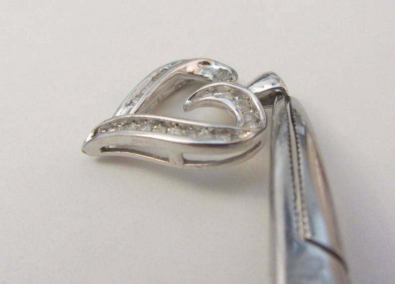 May include: A silver pendant in the shape of a heart, encrusted with small, clear gemstones. The pendant is shown at an angle, with the heart shape slightly open. The metal has a polished, reflective surface.