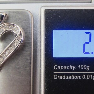 May include: A silver heart-shaped pendant with embedded gemstones, resting on a scale. The scale displays a weight of 2.72 grams. The pendant features a mix of baguette and round-cut stones, creating a sparkling effect.