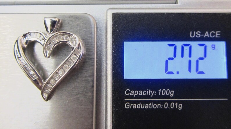 May include: A silver heart-shaped pendant with embedded gemstones, resting on a scale. The scale displays a weight of 2.72 grams. The pendant features a mix of baguette and round-cut stones, creating a sparkling effect.