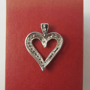 May include: A silver heart-shaped pendant with a halo of small, clear stones. The pendant is set against a red background. The bail is marked "10K %" and "AT".