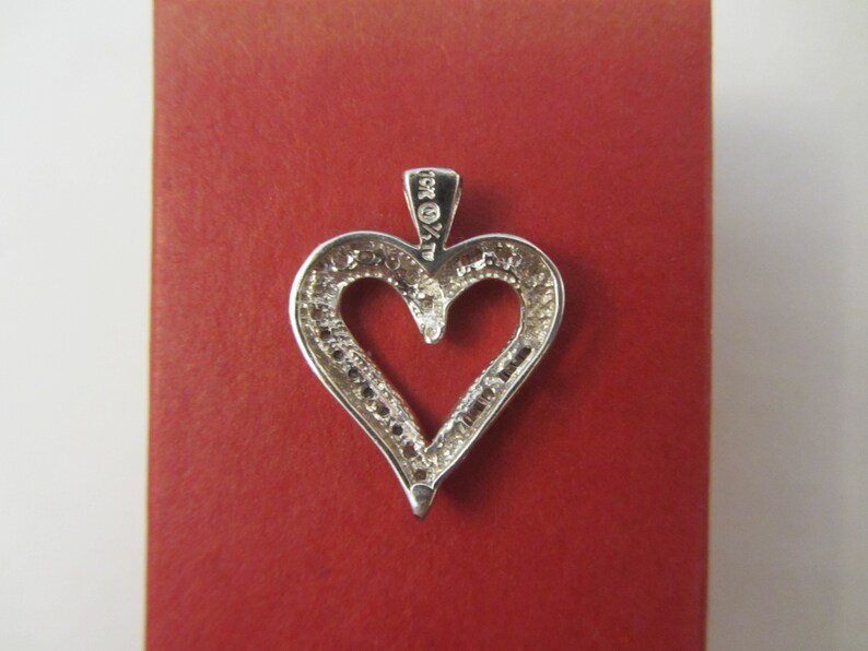 May include: A silver heart-shaped pendant with a halo of small, clear stones. The pendant is set against a red background. The bail is marked "10K %" and "AT".