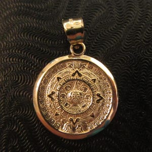 May include: A circular gold pendant featuring a detailed relief of the Aztec sun stone calendar. The design includes a central face surrounded by concentric rings of intricate symbols and geometric patterns, topped with a polished gold bail.