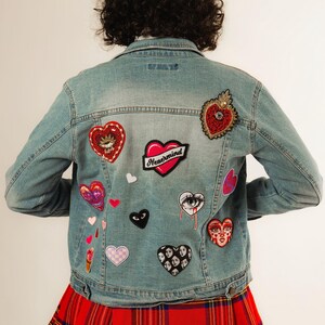 May include: Light blue denim jacket adorned with heart-shaped patches and the word "Nevermind". The jacket is paired with a red plaid skirt. A white baseball cap completes the look, offering a casual, yet stylish aesthetic.
