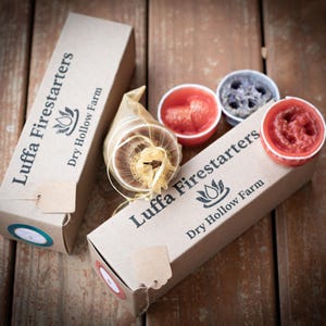 May include: Two boxes of Luffa Firestarters from Dry Hollow Farm. The boxes are brown with black text and a logo. The fire starters are in small, round containers with red, orange, and purple wax.