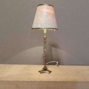 May include: A miniature table lamp with a tall, slender, metallic gold-toned stand featuring decorative rings and a flared base. The lamp has a conical shade in a soft peach hue with a subtle white floral pattern and gold-colored trim at the edges.