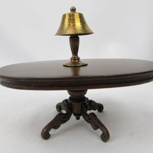 May include: A miniature, dark brown, oval-shaped table with a decorative edge. A small brass lamp with a hammered shade sits on the table. The table has a detailed, carved pedestal base and ornate legs.