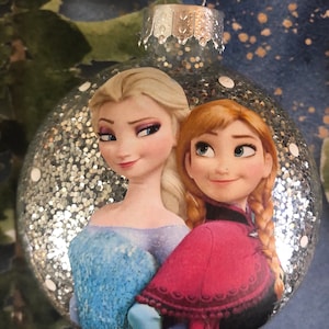 May include: A silver glitter Christmas ornament with a picture of Elsa and Anna from the movie Frozen. The ornament has white polka dots.