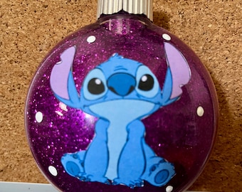 May include: A round, glittery purple Christmas ornament featuring a cartoon image of Stitch from Lilo & Stitch. The ornament has a silver metal cap and a loop for hanging. White dots are scattered across the surface.