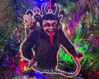 Puede incluir: Un adorno navideño vintage que representa un diablo con una larga lengua roja, sosteniendo una cadena y llevando dos niños a la espalda. El adorno está hecho de madera y tiene una cuerda roja para colgar.