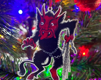 Puede incluir: Un adorno navideño rojo y negro que representa un diablo con una cadena y un látigo. El adorno está colgado de un árbol de Navidad.