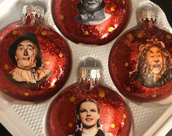 Peut inclure: Ensemble de quatre boules de Noël rouges à paillettes représentant des personnages du Magicien d'Oz. Les boules représentent Dorothy, l'Épouvantail, l'Homme de Fer-Blanc et le Lion peureux.