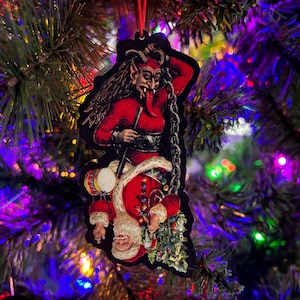 May include: A Christmas ornament featuring a red and black devil holding a chain and a drum, with a Santa Claus figure underneath. The ornament is hanging on a Christmas tree with colorful lights.