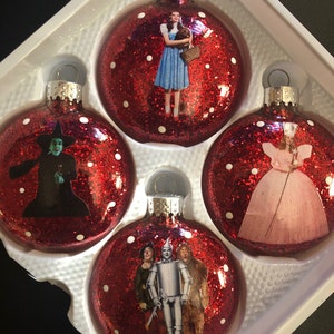 以下が含まれることがあります： オズの魔法使いのキャラクターが描かれた、赤いグリッターのクリスマスオーナメント4個セット。オーナメントには、ドロシー、西の邪悪な魔女、ブリキ男、臆病なライオンが描かれています。