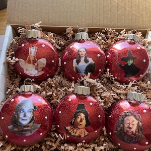 Puede incluir: Seis adornos navideños rojos con purpurina que representan personajes de El Mago de Oz. Cada adorno tiene una imagen diferente, incluyendo a Dorothy, la Bruja Malvada y el Hombre de Hojalata. Los adornos están en una caja de cartón con papel triturado.