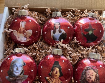 Peut inclure: Six boules de Noël rouges pailletées représentant des personnages du Magicien d'Oz. Chaque boule présente une image différente, dont Dorothy, la méchante sorcière et l'homme de fer-blanc. Les boules sont dans une boîte en carton avec du papier déchiqueté.