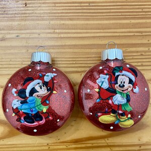 Op de afbeelding: Twee rode glitter kerstballen met afbeeldingen van Mickey Mouse en Minnie Mouse. Beide ballen hebben een rode achtergrond met witte stippen en een kerstverlichting.