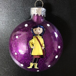 Op de afbeelding: Paarse glitter kerstbal met een wit stippenpatroon. De kerstbal heeft een gedrukt beeld van een meisje dat een gele regenjas en zwarte laarzen draagt.