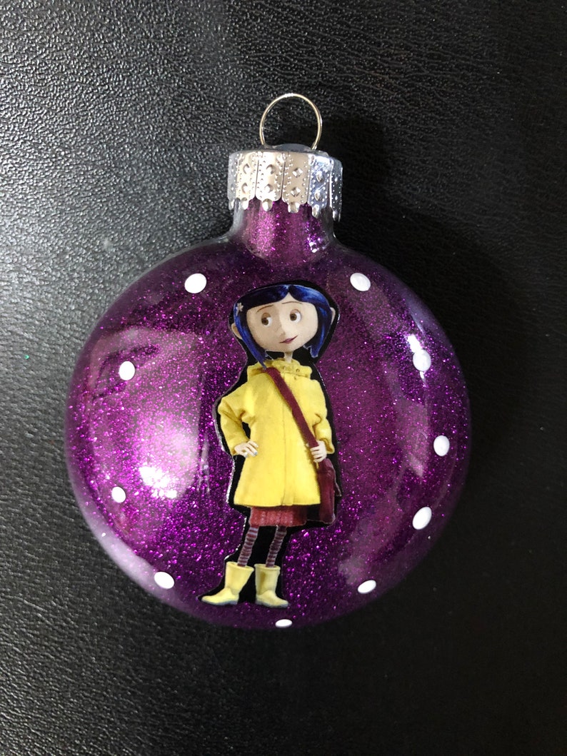 May include: Purple glitter Christmas ornament with a white polka dot pattern. The ornament features a printed image of a girl wearing a yellow raincoat and black boots.