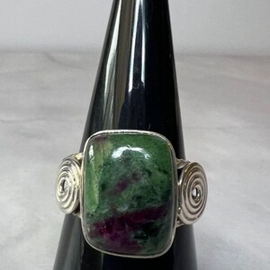 May include: A silver ring with a green and pink gemstone set in a square shape. The ring has two silver swirls on each side of the stone.