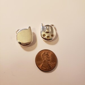 May include: Two silver-toned cat-shaped charms with small loop details. One charm is solid, the other has an "M" design and three yellow gems. A copper penny is next to the charms.