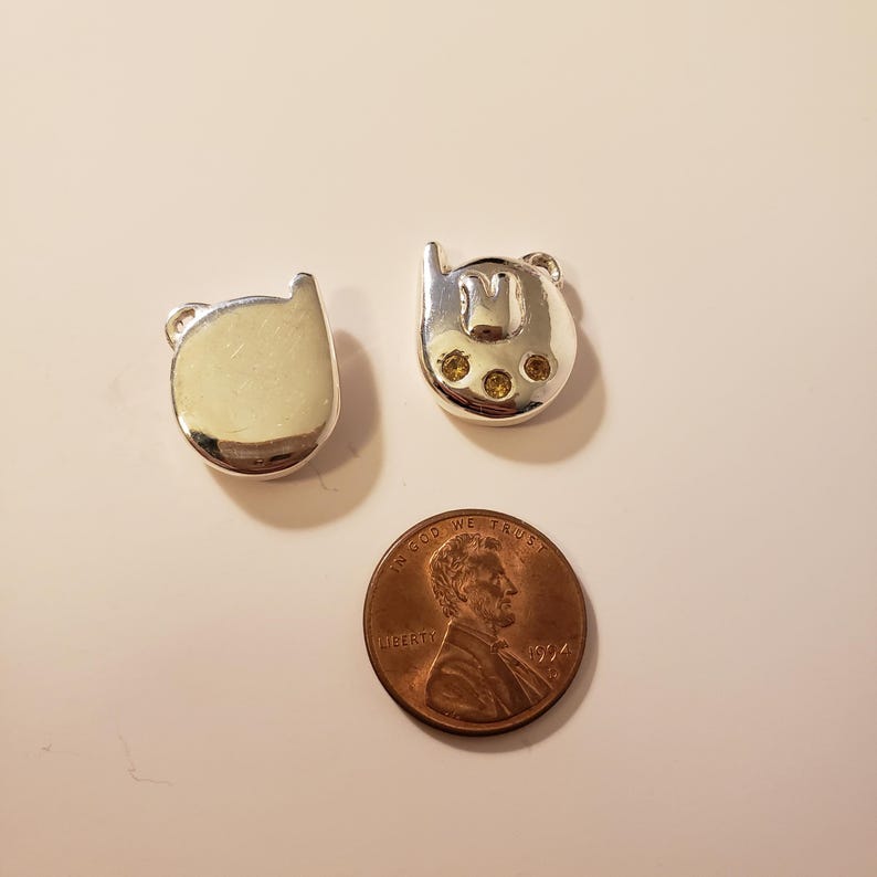 May include: Two silver-toned cat-shaped charms with small loop details. One charm is solid, the other has an "M" design and three yellow gems. A copper penny is next to the charms.