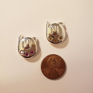 May include: Two silver-toned charms shaped like a stylized letter "M" with small loops at the top. One charm has three yellow gems, the other has three pink gems. A penny is next to the charms.