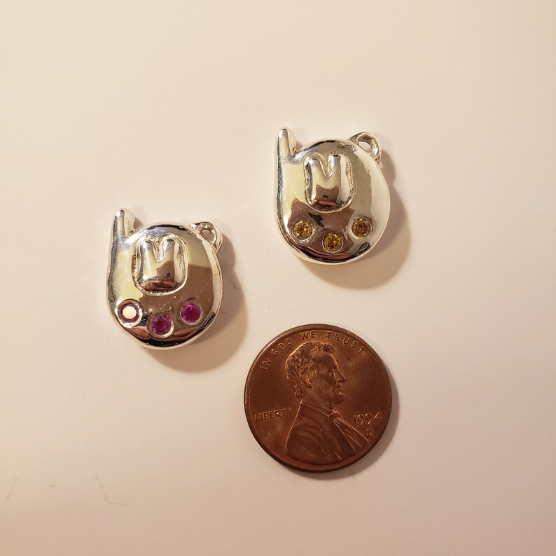 May include: Two silver-toned charms shaped like a stylized letter "M" with small loops at the top. One charm has three yellow gems, the other has three pink gems. A penny is next to the charms.