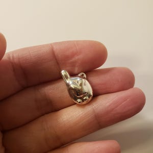 May include: A small, shiny silver charm in the shape of a hand making the "hang loose" gesture. The charm has a loop at the top for attaching to a necklace or bracelet. The hand charm is smooth and reflective.