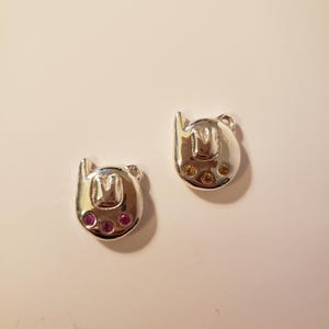 May include: Two silver-toned charms, each shaped like a stylized bear head with the letter "M" in the center. One charm has three pink gems, the other has three yellow gems. The charms are on a white surface.
