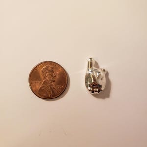 May include: A small, shiny silver charm in the shape of a hand with the index finger pointing up. Next to the charm is a copper penny with the image of Abraham Lincoln. The charm is approximately 0.75 inches in height.