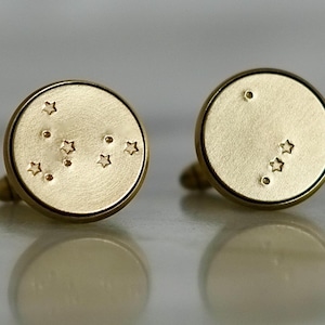 May include: A pair of gold cufflinks with a constellation design. One cufflink features a constellation of stars, while the other cufflink features a single star.