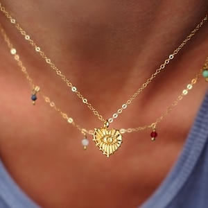 May include: A gold necklace with a heart-shaped pendant featuring an evil eye design. The necklace is layered with a second chain with small colourful beads.