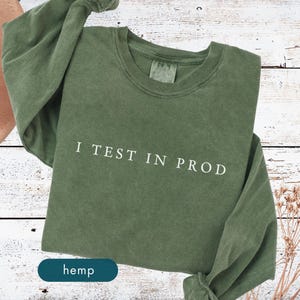 May include: Olive green long-sleeve shirt with the text "I TEST IN PROD" in white. The word "hemp" is in a dark teal rectangle at the bottom. The shirt is on a white wooden surface.
