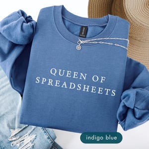 May include: Indigo blue sweatshirt with the text "QUEEN OF SPREADSHEETS" in white. A silver necklace with a pendant rests on the sweatshirt. The image also includes a pair of blue jeans and a straw hat.