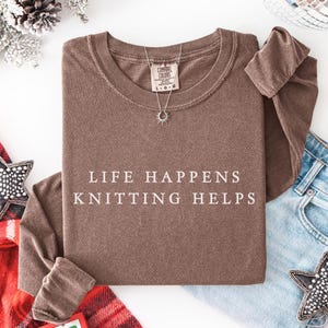 May include: A brown long-sleeve shirt with the text "LIFE HAPPENS KNITTING HELPS" in white. A silver necklace with a sun pendant is on the shirt. The shirt is paired with blue jeans and a decorative star.