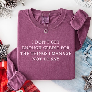 May include: A maroon crew neck sweatshirt with the text "I don't get enough credit for the things I manage not to say." A silver necklace and a pair of blue jeans are next to the sweatshirt. Decorative stars and a plaid scarf are also in the image.
