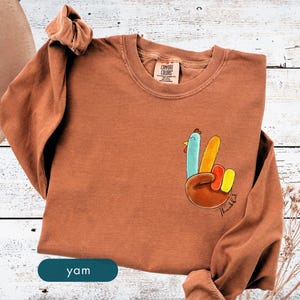 May include: A long-sleeved, rust-colored sweatshirt with a turkey-themed graphic on the front. The graphic depicts a hand making a peace sign, with each finger representing a turkey feather. The word "Thanksful" is written in script.