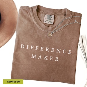 May include: Espresso-colored t-shirt with the words "DIFFERENCE MAKER" in white block letters. A silver necklace with a small pendant rests on the shirt. A portion of a brown hat is visible in the upper left corner.