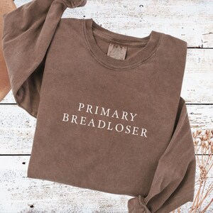 May include: A brown long-sleeve crewneck sweatshirt featuring the text PRIMARY BREADLOSER printed in white across the chest. The garment has a washed, vintage-style appearance with a ribbed collar and cuffs, showcasing a simple, clean design.